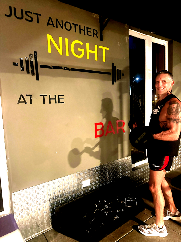 Darren lifting a barbell in front of a sign that reads "Just Another Night at the Bar"